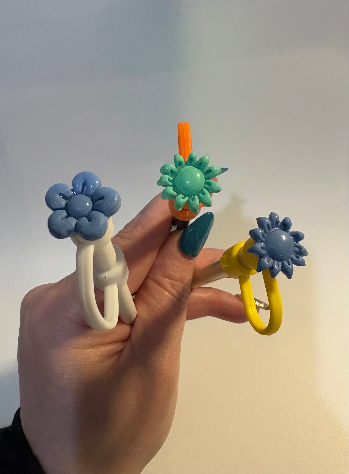Hand holding three colorful handcrafted polymer clay straw cover caps. Unique floral designs in blue and yellow.
