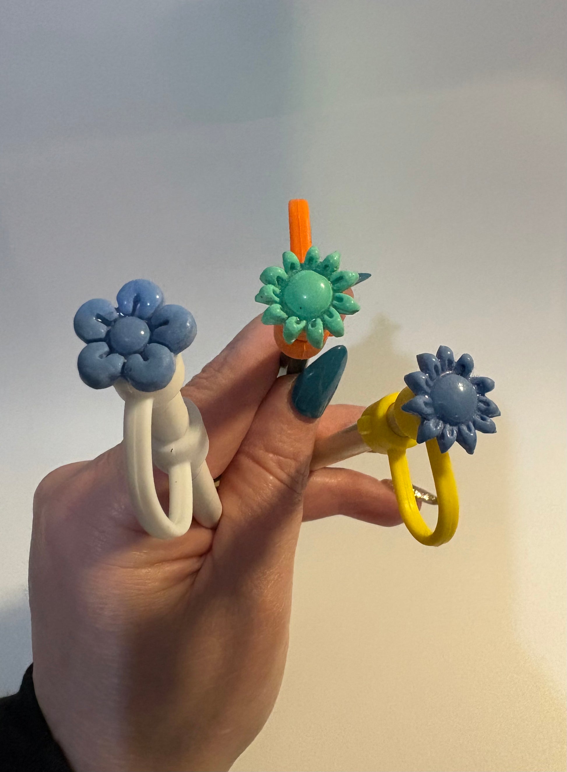 Hand holding three colorful handcrafted polymer clay straw cover caps. Unique floral designs in blue and yellow.